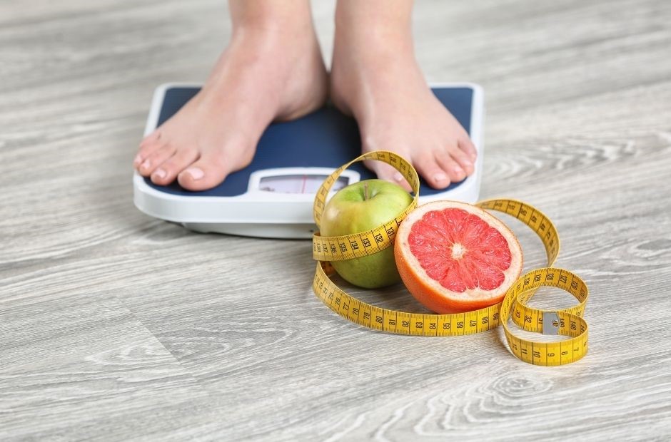 Hollywood-Diät dargestellt von Frauenfüßen auf Waage neben Obst und Maßband